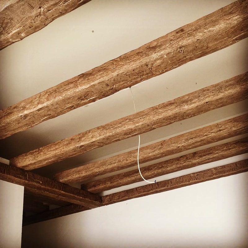 Grinzi din Poliuretan pentru Plafon în Cameră de Oaspeți Grinzile din poliuretan P1981B și căpriorii P1981C reprezintă elemente decorative ce imită fidel textura lemnului natural fasonat, oferind tavanului un aspect rustic și primitor. Când am reamenajat camera de oaspeți, ne-am dorit un spațiu cald în care prietenii și familia să se simtă imediat conectați cu natura. Plafonul simplu și alb din zidărie a fost transformat radical prin adăugarea acestor profiluri dispuse în paralel. Cu o densitate ridicată de 150-220 kg/m³ (țintă ~160 kg/m³), materialul oferă un relief pronunțat cu noduri și fibre lemnoase vizibile, fără a încărca structura de rezistență a clădirii.
Lumina naturală care intră pe fereastră evidențiază fiecare detaliu sculptat al grinzilor din poliuretan, creând un joc fascinant de umbre pe suprafața albă a plafonului. Alegerea acestui sistem decorativ a permis instalarea facilă a cablajului electric suspendat pentru corpurile de iluminat centrale, ascunzând micile imperfecțiuni ale suprafeței de bază. Rezistența excelentă la fluctuații de temperatură între -100 °C / +80 °C garantează că profilurile nu se vor fisura și nu își vor schimba forma în timp, menținând aspectul impecabil al camerei în orice anotimp.
Spre deosebire de grinzile tradiționale din lemn masiv, variantele din poliuretan nu necesită tratamente periodice împotriva cariilor sau barieră suplimentară de protecție la umiditate, având o rată de absorbție a apei de sub 1%. Această proprietate tehnică face ca curățarea zilnică să fie extrem de simplă, fiind suficientă o ștergere ușoară cu o lavetă din microfibră. În plus, greutatea redusă a grinzilor P1981B a permis montajul rapid doar prin lipire cu adeziv structural dedicat, fără a fi nevoie de șuruburi masive sau sprijin mecanic complex.
Rezultatul final în camera de oaspeți este o atmosferă echilibrată, unde căldura vizuală a tonurilor de maro nuc se îmbină perfect cu minimalismul pereților albi. Fiecare musafir care ne trece pragul admiră naturalețea tavanului și este surprins să afle că nu este vorba despre un lemn greu de tăiat, ci despre o soluție modernă și practică din poliuretan. Integrarea grinzilor decorative P1981B și a mertecilor P1981C transformă o încăpere obișnuită într-un spațiu memorabil, plin de personalitate și confort.