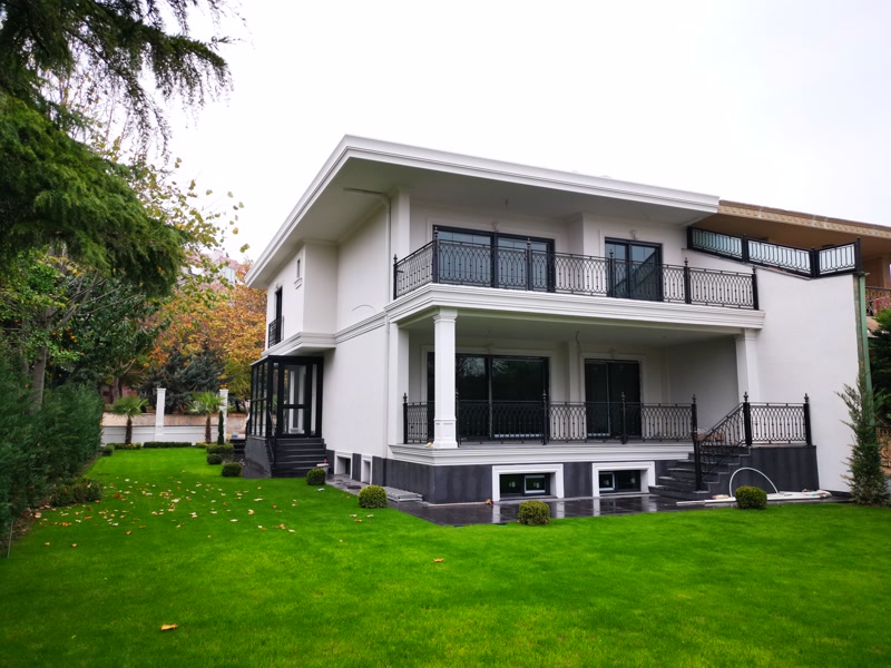 Profile Exterioare din Poliuretan pentru Fațadă de Vilă Decorarea fațadei unei vile cu profile din poliuretan de înaltă densitate (aproximativ 150-220 kg/m³ (țintă ~160 kg/m³)) reprezintă o metodă excelentă pentru a obține o estetică arhitecturală deosebită și o izolație vizuală impecabilă. Când am decis să renovăm exteriorul casei noastre, dorința noastră principală a fost să creăm un spațiu călduros, potrivit pentru viața de familie și momentele de relaxare în aer liber. Fiecare zi petrecută în această vilă începe cu bucuria de a privi armonia dintre grădina verde și albul curat al pereților. Profilele exterioare din poliuretan au fost alegerea perfectă datorită rezistenței lor extraordinare la variații extreme de temperatură cuprinse între -100 °C / +80 °C.
Zona terasei principale și balconul de la etaj sunt susținute vizual de pilatrii pătrați din poliuretan cod P5030 și bazele decorative P5030B. Aceste elemente verticale oferă structurii o proporție echilibrată și creează o zonă umbrită ideală pentru cinele în familie sau serile de lectură. Poliuretanul de înaltă calitate utilizat are un coeficient de absorbție a apei de sub 0.1%, ceea ce previne infiltrările și degradarea cauzată de ploaie sau zăpadă. Pe terasa exterioară, aceste coloane adaugă o notă de eleganță sobră, făcând ca spațiul să devină extensia naturală a livingului.
Pentru conturarea ferestrelor mari și a ușilor glisante am ales ancadramente din poliuretan din gamele P1454, P1462, P1465 și P9001. Aceste profile decorative creează rame elegante în jurul suprafețelor vitrate, sporind senzația de volum și oferind o personalitate unică întregii clădiri. Ferestrele mari permit luminii soarelui să lumineze interiorul vilei, în timp ce ancadramentele exterioare conferă un aspect îngrijit și structurat. După montarea cu adeziv special, am aplicat 2 straturi de vopsea lavabilă de exterior pe bază de apă, asigurând o protecție durabilă împotriva razelor ultraviolete.
Rezultatul final este o casă modernă, senină și plină de viață, unde contrastele dintre albul fațadei, feroneria neagră și verdele curții oferă o stare permanentă de confort. Un exterior amenajat cu atenție la detalii transformă simpla locuință într-un cămin autentic și primitor pentru toți membrii familiei. Cu ajutorul soluțiilor arhitecturale de la Decoratiuni Casa, am reușit să îmbinăm durabilitatea materialelor moderne cu frumusețea unui stil de viață relaxat și primitor.