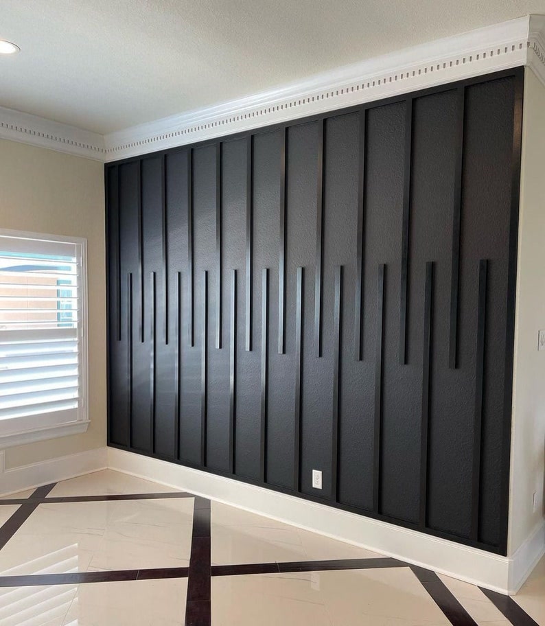 Perete Accent Negru cu Profil Poliuretan Decorativ Poliuretanul profil decorativ P851350 este un element arhitectural din spumă poliuretanică cu densitate de 150-220 kg/m³ (țintă ~160 kg/m³), utilizat pentru crearea reliefurilor geometrice pe pereții interiori. În livingul nostru primitor, am transformat peretele principal dintr-o suprafață simplă într-o zonă centrală de interes prin aplicarea acestui profil într-un model vertical asimetric. Nuanța mată de negru antrasit oferă o profunzime vizuală deosebită, contrastând elegant cu finisajele deschise ale spațiului. Lumina naturală care pătrunde prin ferestrele cu obloane albe creează jocuri de umbre și lumini pe suprafața texturată, aducând un plus de căldură și dinamism în fiecare zi.
Experiența de zi cu zi în această zonă de zi este complet schimbată de prezența peretelui de accent. Datorită rezistenței poliuretanului la variații de temperatură între -100 °C / +80 °C și a gradului de absorbție a apei de sub 1%, structura își păstrează stabilitatea dimensională fără fisuri în timp. Am combinat liniile verticale drepte ale profilelor subțiri cu o plintă albă generoasă la bază și o scofie clasică profilată la îmbinarea cu tavanul. Această juxtapunere între stilul contemporan al baghetelor negre și elementele clasice albe oferă sufrageriei un caracter rafinat, fiind cadrul perfect pentru întâlnirile de familie sau momentele de relaxare de seară.
Procesul de montaj al profilelor din poliuretan P851350 s-a dovedit extrem de simplu și curat pentru locuința noastră. Baghetele au fost debitate la lungimi variabile cu un ferăstrău manual, iar după o ușoară șlefuire cu hârtie abrazivă cu granulație 220, au fost fixate direct pe perete utilizând un adeziv acrilic de înaltă rezistență. După montare, suprafața a fost protejată cu 2 straturi de vopsea lavabilă pe bază de apă într-o nuanță închisă de cărbune. Această metodă de decorare aduce volum fără a încărca spațiul util, transformând sufrageria într-un mediu primitor și confortabil.
Pardoseala din plăci ceramice de mari dimensiuni cu inserții geometrice de culoare închisă completează perfect compoziția peretelui decorativ. Modul în care reflexiile luminii pe suprafața lucioasă a pardoselii interacționează cu textura mată a peretelui accent creează un echilibru vizual plăcut. Alegerea profilelor din poliuretan permite o personalizare nelimitată a spațiilor de locuit, oferind familiilor posibilitatea de a-și exprima stilul propriu într-un mod durabil și estetic.