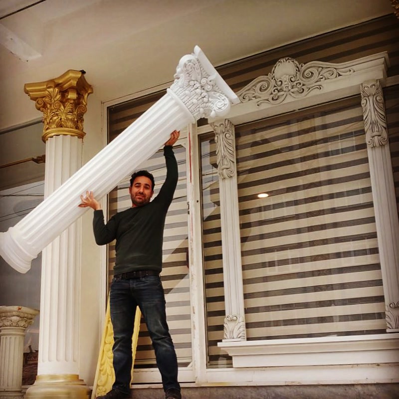 Un bărbat susține cu ușurință o coloană decorativă albă din poliuretan în fața unei ferestre ornamentate.