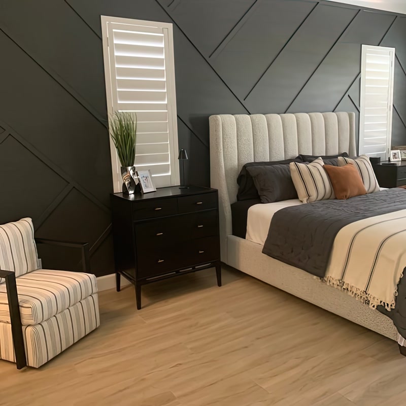 Ancadramente din Poliuretan pentru Perete Accent în Dormitor Bagheta din poliuretan P851350 este un element decorativ de înaltă densitate conceput pentru a crea modele geometrice dinamice pe suprafețele verticale interioare. Diminețile în acest dormitor reamenajat încep cu o senzație plăcută de liniște și confort vizual, unde peratele accent devine piesa centrală a spațiului. Nuanța închisă de antracit oferă o profunzime deosebită spațiului de odihnă, iar lumina naturală ce pătrunde prin jaluzelele albe evidențiază fiecare linie decorativă, creând un joc subtil de umbre și lumini pe parcursul întregii zile.

Din punct de vedere tehnic, realizarea acestui model geometric asimetric pe perete implică debitarea profilelor la unghiuri precise de 45 de grade și fixarea lor cu adeziv special. Profilele din poliuretan au o densitate ridicată de aproximativ 150-220 kg/m³ (țintă ~160 kg/m³) și rezistă excelent la umiditate, având o rată de absorbție a apei de sub 1%. Datorită rezistenței termice în intervalul cuprins între -100 °C / +80 °C, materialul își păstrează stabilitatea dimensională în timp și menține îmbinările perfect liniare. După montaj, întreaga suprafață a fost acoperită uniform cu 2 straturi de vopsea lavabilă mată pe bază de apă.

Senzația de căldură din încăpere este completată armonios de parchetul din lemn în tonuri calde de stejar și de patul tapițat cu tăblie înaltă. Mobilierul de culoare închisă și fotoliul confortabil cu imprimeu în dungi creează un colț intim ideal pentru lectură în serile liniștite. Textilele alese în nuanțe neutre de gri, crem și accente de teracotă echilibrează tonul închis al fundalului, transformând dormitorul într-un refugiu primitor pentru întreaga familie.

Utilizarea profilului din poliuretan P851350 permite adaptarea ușoară a designului în funcție de dimensiunile fiecărei camere. Proprietarii pot alege compoziții simetrice sau structuri diagonale libere pentru a personaliza spațiul fără a încărca vizual decorul. Întreținerea suprafețelor decorate se realizează extrem de simplu, prin ștergere periodică cu o lavetă din microfibră ușor umezită, ceea ce face ca această soluție decorativă să fie deosebit de practică pentru un stil de viață modern.