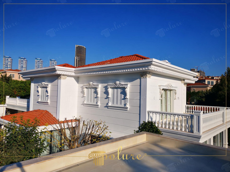 Fațade exterioare cu măreție arhitecturală clasică, îmbogățite cu coloane și detalii din poliuretan Eleganța arhitecturii clasice este evidentă și în clădirile moderne. Vila din această imagine prezintă unul dintre cele mai frumoase exemple de decorare exterioară clasică. Nobilimea albului intalneste coloanele din poliuretan si capitelurile de coloana in stil corintic. În timp ce stâlpii ferestrelor adaugă profunzime detaliilor elegante de pe fațadele exterioare, badastrolul și mulurile de streașină fac silueta caracteristică a clădirii clară. Acoperiri cu panouri din poliuretan Aduce frumusețe de lungă durată fațadelor exterioare, îmbinând estetica tradițională cu avantajele materialelor moderne. Fie că proiectați o clădire nouă sau vă gândiți să adăugați o notă clasică unei case existente, aceste elemente adaugă originalitate și valoare estetică fiecărui proiect.