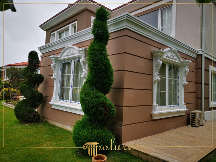 Decorații estetice de fațadă din poliuretan care ies în evidență printre verde Această imagine demonstrează perfect estetica și funcționalitatea poliuretanului în decorațiunile exterioare clasice. Pereții în tonuri de maro deschis sunt însuflețiți cu stâlpi din poliuretan alb, muluri de streașină și decorațiuni superioare în formă de coroană și contraforturi elegante completează designul ferestrei estetica generală a clădirii. Acoperirile cu panouri din poliuretan se remarcă prin durabilitate și se adaugă la fațada exterioară. Acest design personaliza și îmbogățește exteriorul fiecărei locuințe combinând elementele clasice și moderne într-un mediu înconjurat de verdeață.